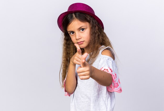 confident little caucasian girl with purple party hat pointing  isolated on white wall with copy space