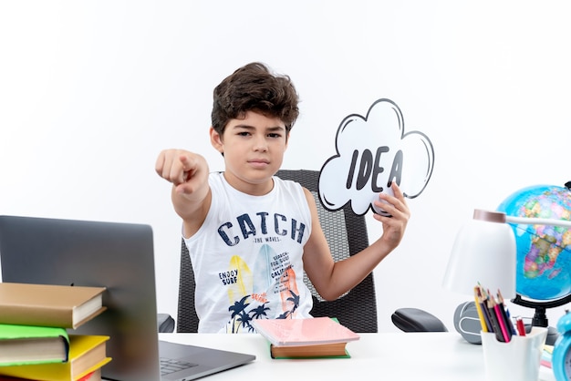 Confident little schoolboy sitting at desk with school tools holding idea bubble and showing you gesture isolated on white background