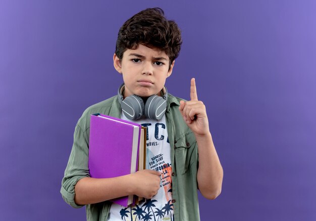 Confident little schoolboy wearing headphones holding books and points at up 