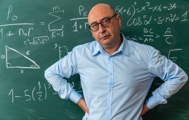 Confident looking camera middle-aged male teacher wearing glasses standing in front blackboard putting hands on hip