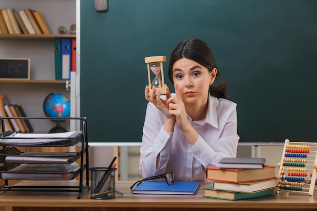 教室で学校のツールと机に座って砂時計を保持しているカメラの若い女性教師を見て自信を持って