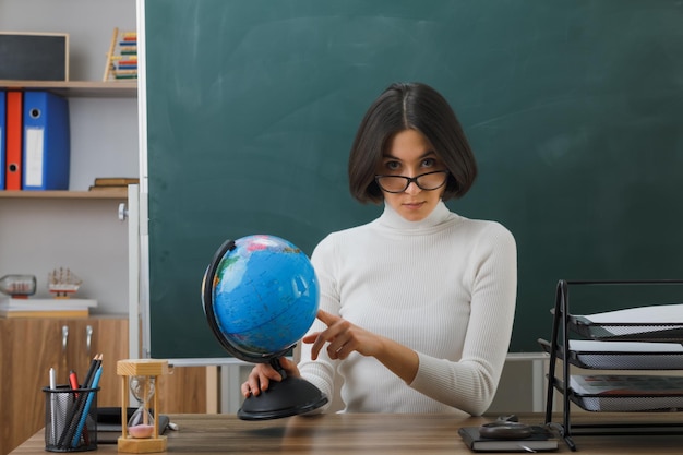 confident looking at camera young female teacher wearing glasses holding and points at world globe sitting at desk with school tools on in classroom