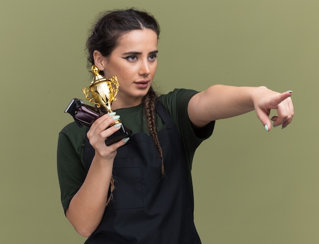 Confident looking at side young female barber in uniform holding hair clippers with winner cup points at side isolated on olive green wall
