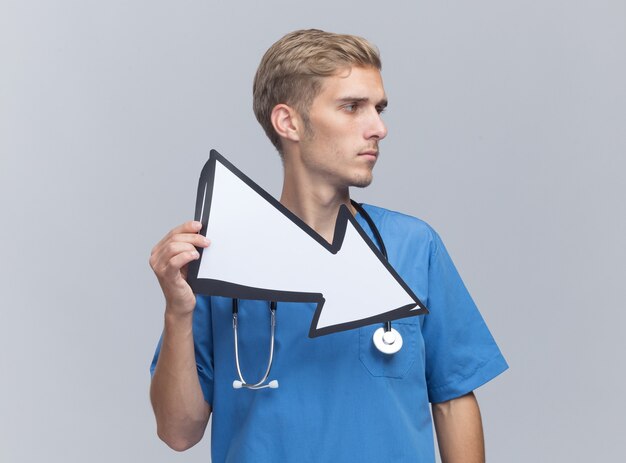 Confident looking at side young male doctor wearing doctor uniform with stethoscope holding direction mark isolated on white wall