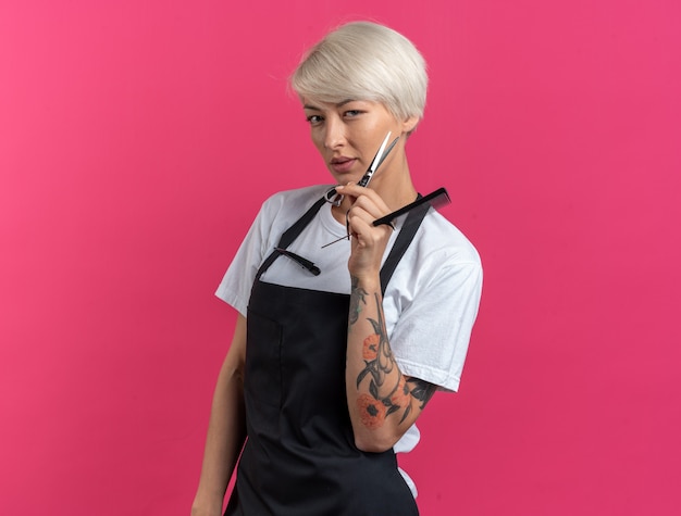 Бесплатное фото Уверенно выглядящая молодая красивая женщина-парикмахер в униформе, держащая ножницы с расческой, изолированной на розовой стене