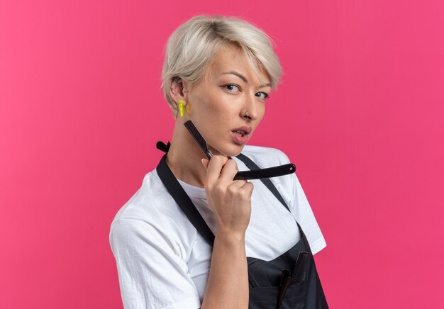 Confident looking  young beautiful female barber in uniform holding straight razor isolated on pink wall