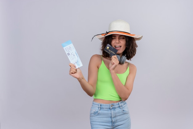 Уверенная в себе милая молодая женщина с короткими волосами в зеленом топе в шляпе от солнца показывает билеты на самолет и кредитную карту на белом фоне