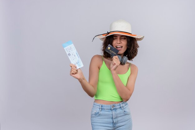 A confident lovely young woman with short hair in green crop top wearing sun hat showing plane tickets and credit card on a white background