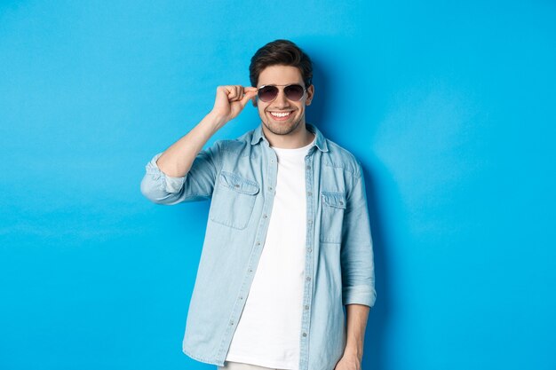 Confident macho man put on sunglasses, looking cool and sassy, standing over blue background