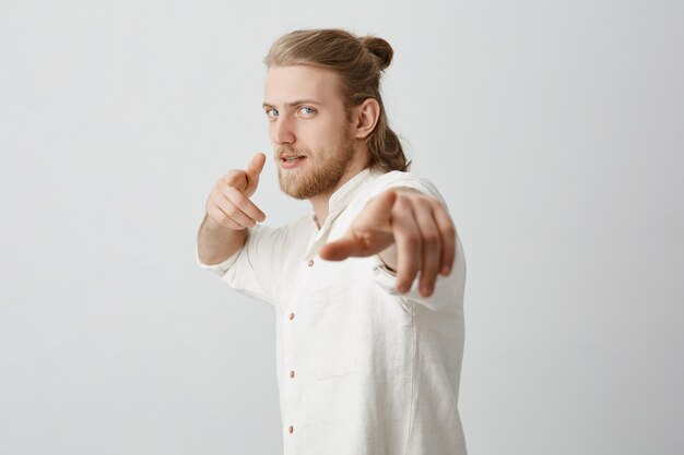 Confident macho man with blond hair and bun, pointing at camera with index fingers,