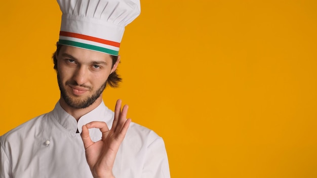 주황색 배경 위에 OK 사인을 보여주는 광고 공간 근처에 제복을 입은 자신감 있는 남성 요리사 승인된 제스처를 보여주는 요리사 모자를 쓴 남자