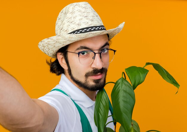 Confident male gardener in optical glasses wearing gardening hat holds plant and pretends to hold camera 