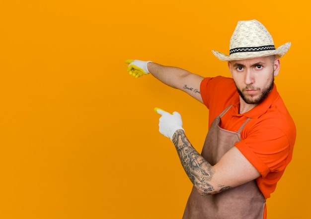 Confident male gardener wearing gardening hat and gloves points at back with two hands looking  