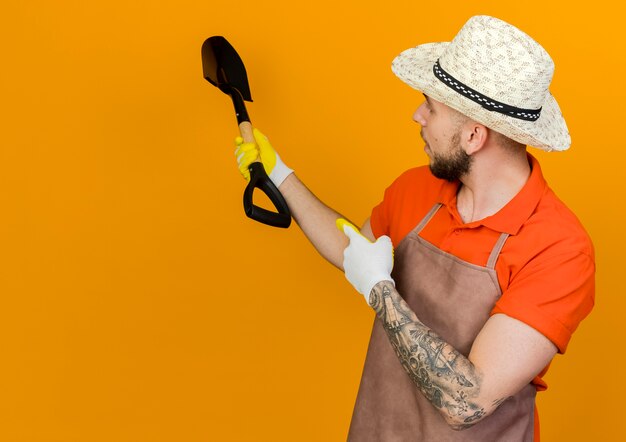 Confident male gardener wearing gardening hat holds spade looking and pointing behind 