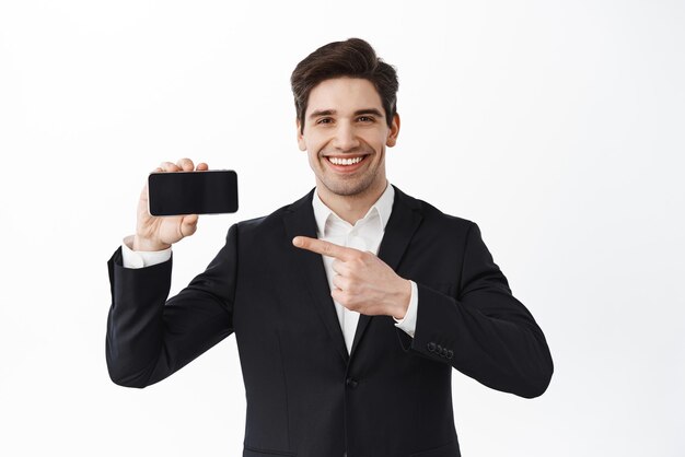 Confident male office worker ceo manager pointing at horizontal phone screen and smiling demonstrate website or application standing in suit against white background