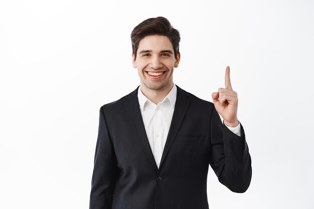 Confident male real estate agent, broker business man in suit pointing finger up, showing top advertisement, deal on market, standing over white background