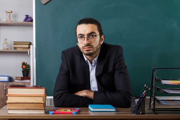 Confident male teacher wearing glasses sitting at table with school tools in classroom