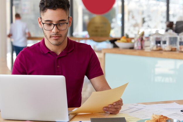 Confident man in casual outfit, provides electronic payment, uses online banking application on laptop computer, develops website page, holds papers, works in cozy coffee shop. Modern technologies