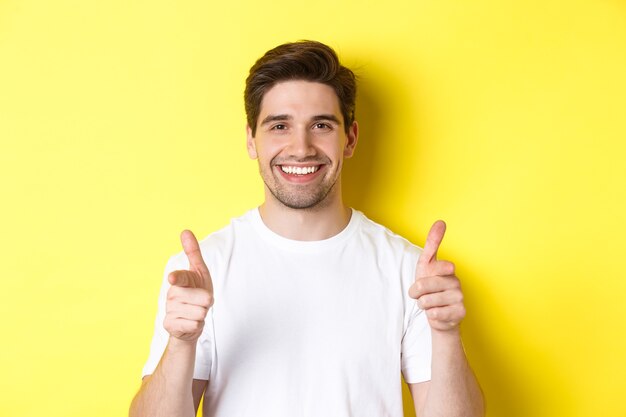 Confident man pointing fingers at camera and smiling, praising you, standing over yellow background.