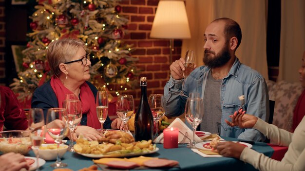 크리스마스 저녁 식사에서 스파클링 와인을 마시는 동안 긍정적인 고위 여자와 이야기 하는 자신감 남자. 전통 음식과 샴페인으로 겨울 축제를 축하하는 쾌활한 다인종 가족.