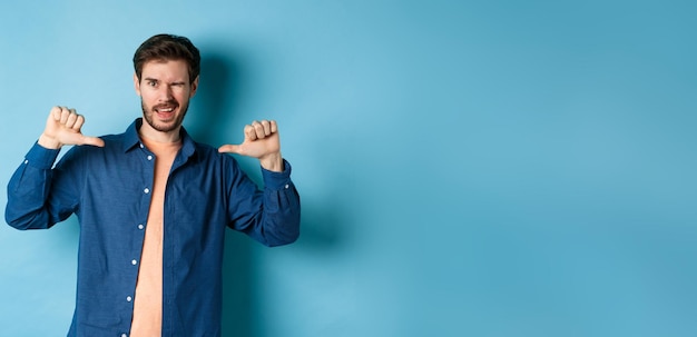 Free photo confident man winking and pointing at himself selfpromoting standing on blue background