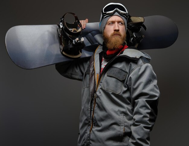 Confident man with a red beard wearing a full equipment holding a snowboard on his shoulder, looking away with a serious look