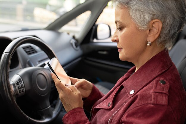 Confident mature businesswoman in stylish jacket chats with her smartphone inside the car