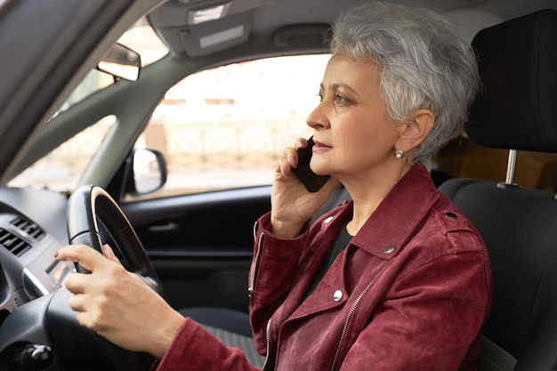 Fiduciosa donna di affari matura in giacca elegante alla guida di auto per le strade della città e parlando al cellulare contemporaneamente