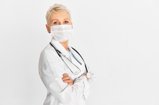 Confident mature woman physician in protective medical mask and uniform crossing arms on her chest posing against blank studio wall background with copy space for your advertising information