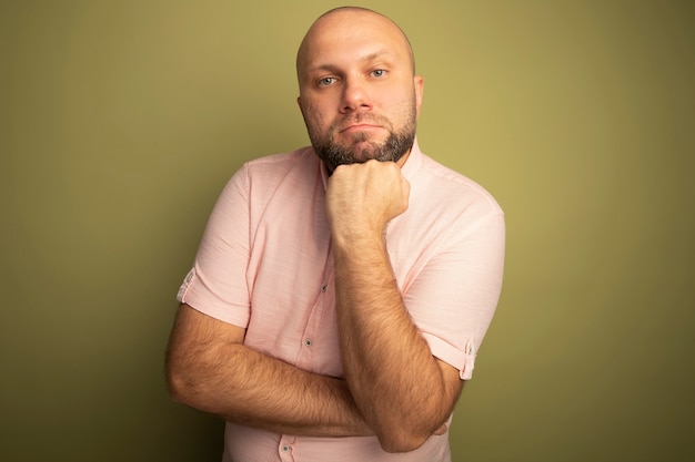 Уверенный в себе лысый мужчина средних лет в розовой футболке, положив руку под подбородок на оливково-зеленом фоне