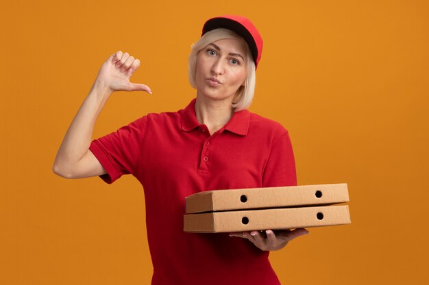 Confident middle-aged blonde delivery woman in red uniform and cap holding pizza packages looking at front pointing at herself with pursed lips isolated on orange wall