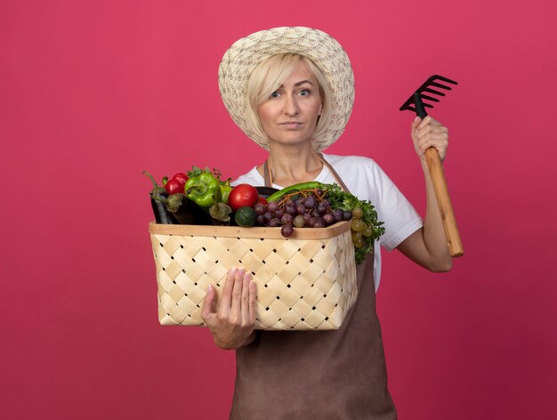Уверенная блондинка среднего возраста женщина-садовник в униформе в шляпе держит корзину с овощами и граблями, глядя вперед, изолированную на малиновой стене с копией пространства