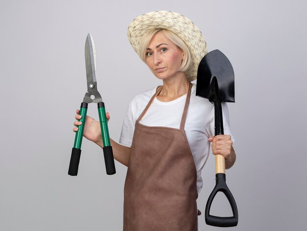Confident middle-aged blonde gardener woman in uniform wearing hat holding hedge shears and spade