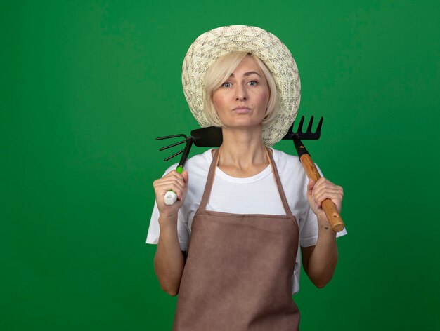 Confident middle-aged blonde gardener woman in uniform wearing hat holding rake and hoe-rake on shoulders