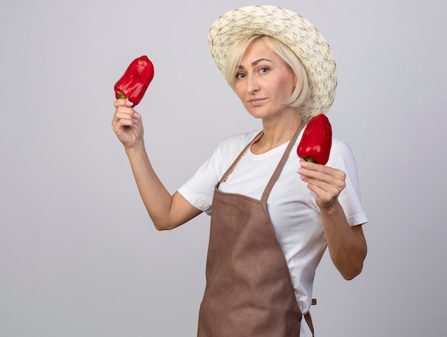 Confident middle-aged blonde gardener woman in uniform wearing hat standing in profile view holding peppers  isolated on white wall