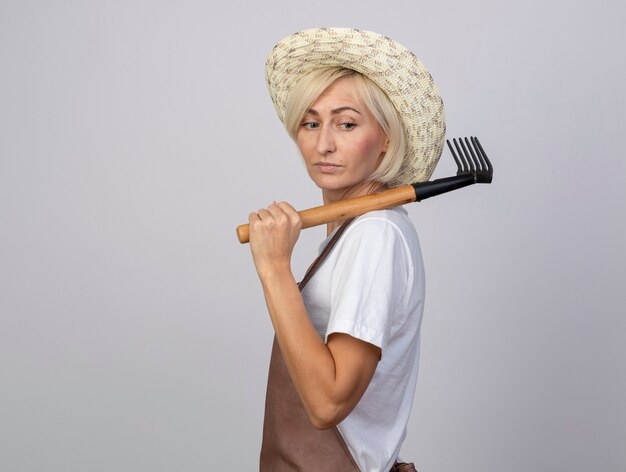 Confident middle-aged blonde gardener woman in uniform wearing hat standing in profile view holding rake on shoulder looking at side