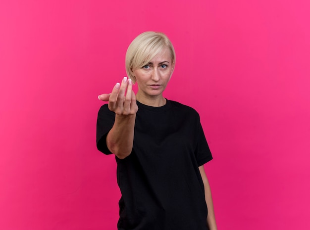 Confident middle-aged blonde slavic woman  doing come here gesture isolated on crimson wall with copy space