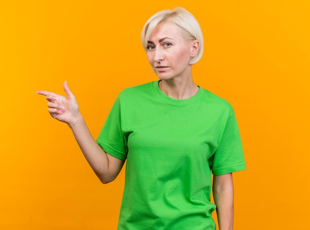 Free photo confident middle-aged blonde slavic woman looking at camera pointing at side isolated on yellow background with copy space