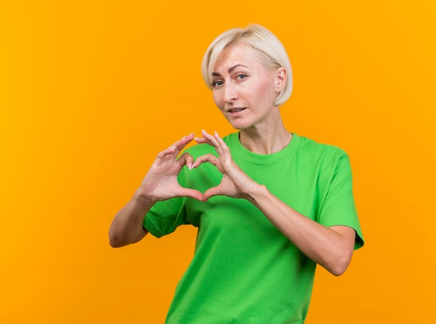 Fiduciosa donna slava bionda di mezza età guardando la parte anteriore facendo segno di cuore isolato sulla parete gialla con lo spazio della copia