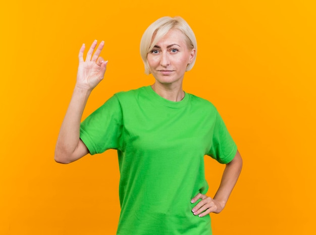Confident middle-aged blonde slavic woman looking at front keeping hand on waist doing ok sign isolated on yellow wall with copy space