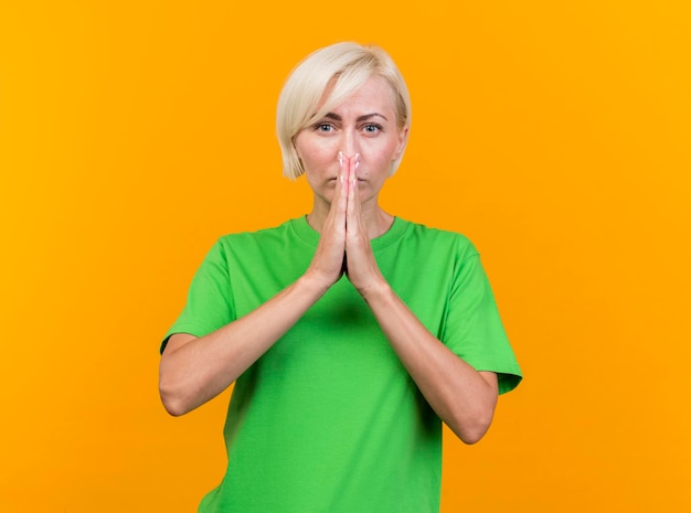 Free photo confident middle-aged blonde slavic woman looking at front keeping hands together in pray gesture in front of mouth isolated on yellow wall with copy space