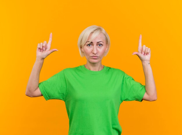 Confident middle-aged blonde slavic woman looking at front pointing up isolated on yellow wall