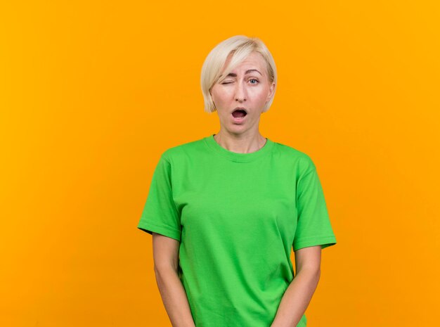 Confident middle-aged blonde slavic woman looking at front and winking isolated on yellow wall with copy space