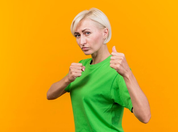 Confident middle-aged blonde slavic woman standing in profile view looking at front doing boxing gesture isolated on yellow wall with copy space