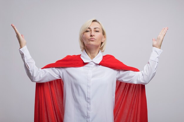 Confident middle-aged blonde superhero woman in red cape  keeping hands in air isolated on white wall