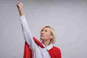 Free photo confident middle-aged blonde superhero woman in red cape raising fist up looking at it