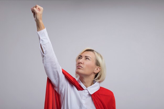 Confident middle-aged blonde superhero woman in red cape raising fist up looking at it