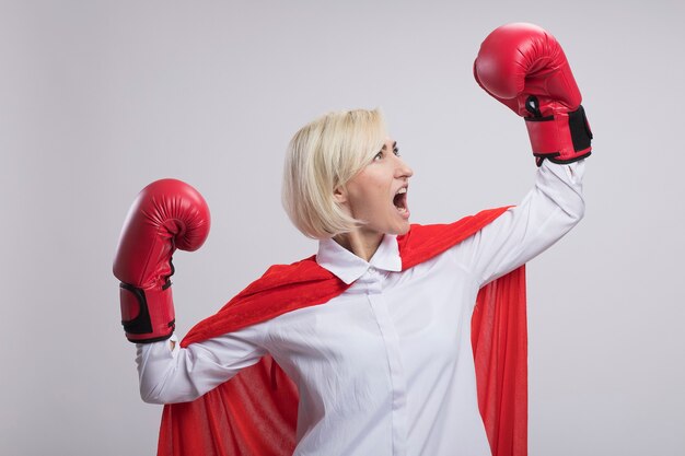 Confident middle-aged blonde superhero woman in red cape wearing box gloves doing strong gesture looking at her fist screaming isolated on white wall