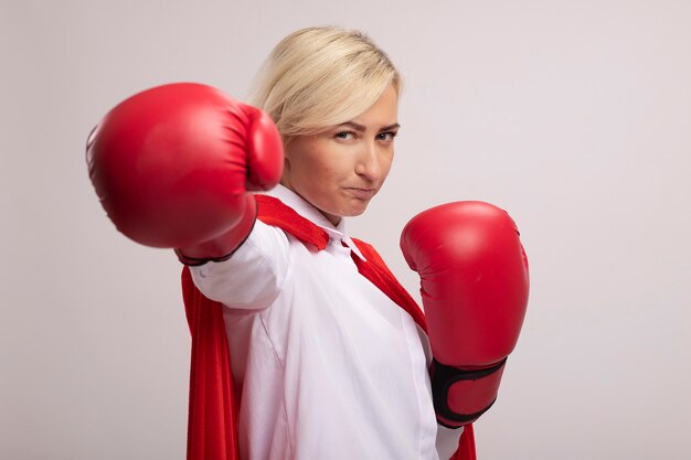 Confident middle-aged blonde superhero woman in red cape wearing box gloves stretching out fist towards camera 