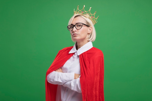 Foto gratuita fiduciosa bionda di mezza età supereroe donna in mantello rosso con gli occhiali e la corona in piedi con postura chiusa in vista di profilo